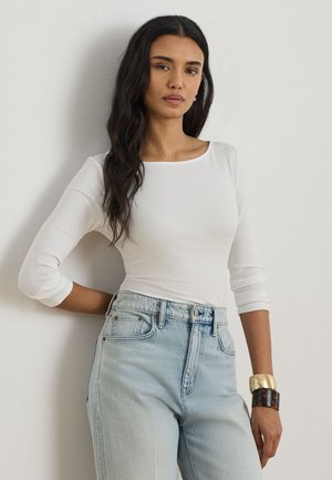 Ženska z dolgimi temnimi lasmi, ki nosi oprijeto belo majico z dolgimi rokavi, svetlomodre kavbojke z visokim pasom in dve masivni zapestnici.