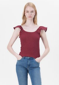 Burgundy top with a smocked texture, featuring ruffled lace sleeves. Model wears blue high-waisted jeans, standing with hands in pockets.