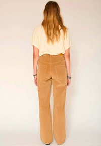 Pantalons en velours côtelé à jambes larges de couleur brun clair avec une finition texturée et deux poches arrière. Associés à un haut crème.