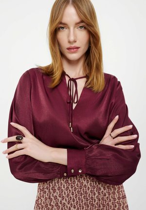 Blusa in raso bordeaux con scollatura a V e fiocco, maniche lunghe a palloncino con bottoni decorativi, abbinata a una gonna con motivo.