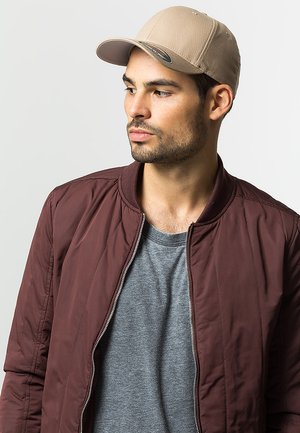 Tan pet met textuurstof en een gebogen klep, gecombineerd met een bordeauxrode bomberjack en een grijs t-shirt. Neutrale achtergrond.