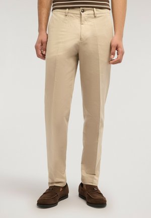 Mann trägt beige maßgeschneiderte Hose, braune Wildleder-Loafer und ein braun gestreiftes Hemd, steht vor einem neutralen Hintergrund.
