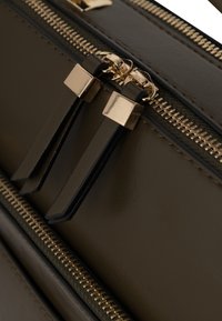 Zielona oliwkowa skórzana torba w zbliżeniu, z złotymi zamkami błyskawicznymi i czarnymi akcentami. Do zauważalnych szczegółów należą podwójne uchwyty zamków o prostokątnych końcach.