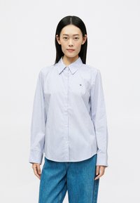 Žena s rovnými čiernymi vlasmi má oblečenú svetlomodrú košeľu s vertikálnymi pruhmi a voľné modré džínsy, stojaca na pozadí z bielej plochy.