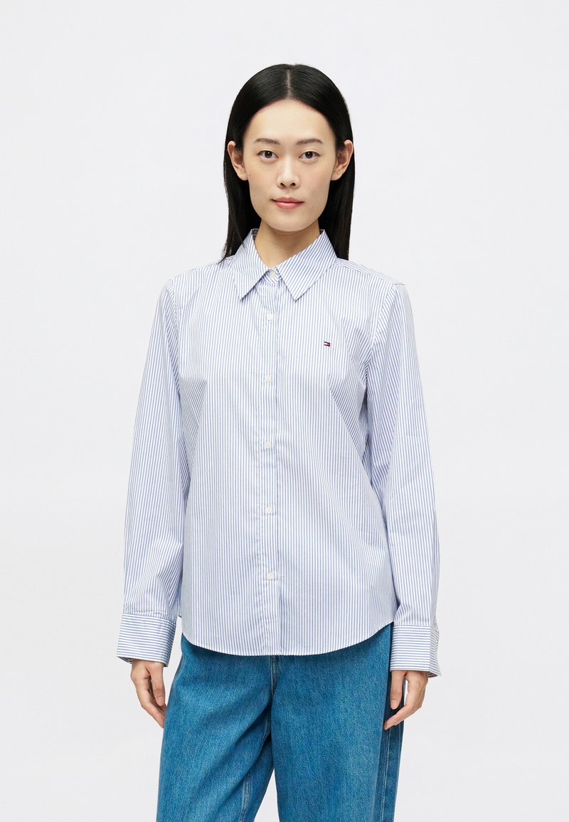 Žena s rovnými čiernymi vlasmi má oblečenú svetlomodrú košeľu s vertikálnymi pruhmi a voľné modré džínsy, stojaca na pozadí z bielej plochy.