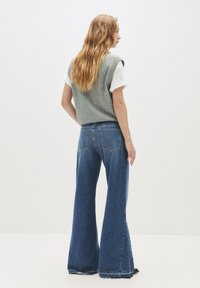 Mladá žena s dlhými vlnitými vlasmi, oblečená v šedom bezrukávovom svetre cez biele tričko s vyhrnutými rukávmi a širokými modrými džínsoch, stojí bokom.