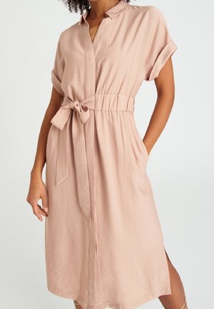 Vrouw draagt een lichtroze, korte mouwen, midi-jurk met kraag, een ceintuur in de taille en zijzakken.