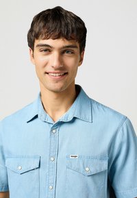 Camicia di jeans azzurro chiaro a maniche corte con bottoni a pressione, due tasche sul petto e un'etichetta con il logo Wrangler sulla tasca sinistra.