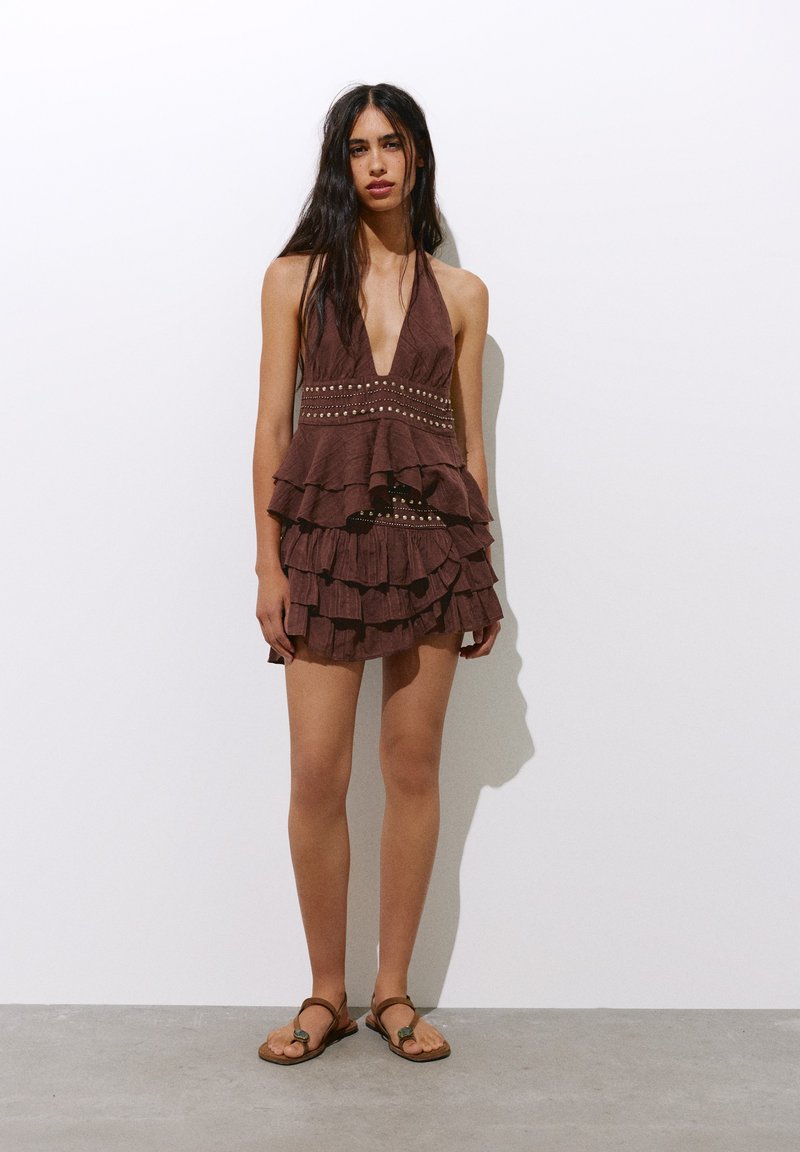 Femme debout contre un mur blanc portant une mini robe marron sans manches à volants avec une taille cloutée et des sandales plates marron.
