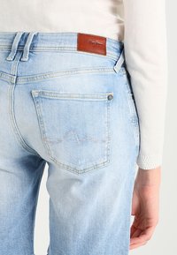 Jeans de denim azul claro con un lavado medio, que cuentan con un parche de cuero marrón, bolsillos estándar y un detalle de costura sutil en el bolsillo trasero.