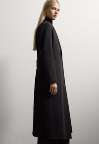 Blonde vrouw in een zwarte lange wollen jas en zwarte coltrui, staand in profiel tegen een effen witte muur.
