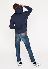 Muž v námořnickém mikině s kapucí, s ohrnutými modrými džíny, černými ponožkami a bílými teniskami, otočený zády s jednou rukou dotýkající se hlavy.