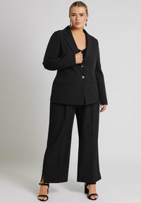 Svart skräddarsydd kostym bestående av en kavaj med djup V-ringning, två framfickor och silverknappar, tillsammans med vida byxor.