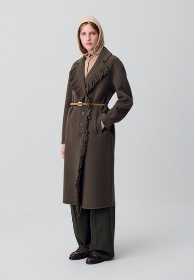 Cappotto di lana marrone con dettagli a frange lungo il colletto, chiuso con una cintura. Abbinato a pantaloni a gamba larga olive e scarpe scure.