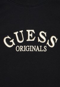 Svart sweatshirt med texten "GUESS" och "ORIGINALS" i broderad gräddfärgad lettering. Slät tyg med ribbad halsringning.