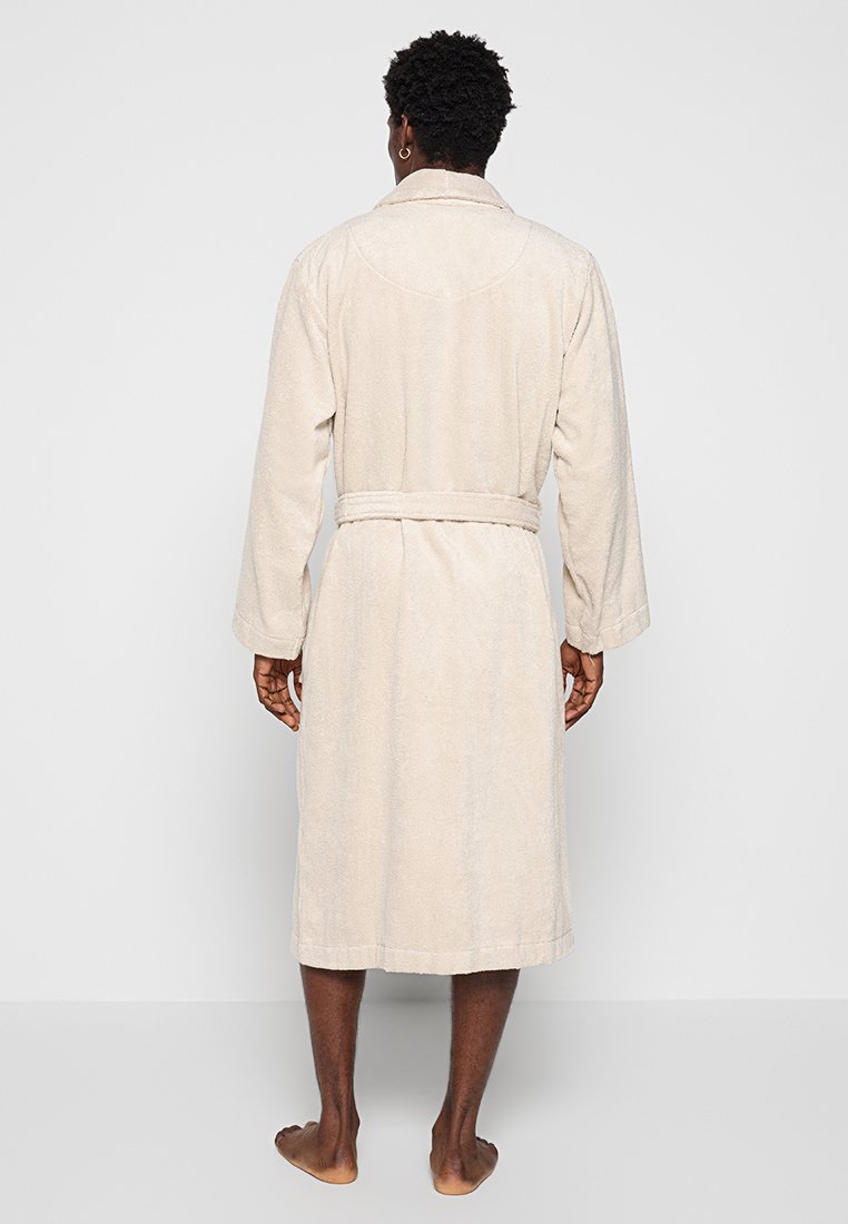 Mężczyzna stojący boso, odwrócony plecami, ubrany w beżowy szlafrok do kolan z paskiem związanym w talii, na jednolitym jasnym tle.