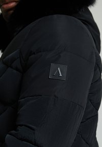 Giacca a vento nera con un morbido colletto in pelliccia, texture trapuntata e una patch con logo in gomma sulla spalla. Tutti i materiali sono morbidi.