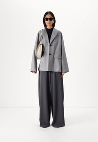 Grå oversized blazer med to sorte knapper og sidelommer, parret med vide mørkegrå bukser og en lys grå håndtaske.