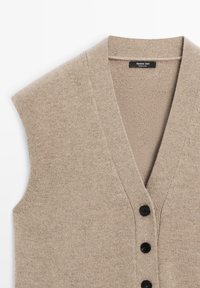 Beżowa dzianinowa kamizelka z dekoltem w serek, z czarnymi guzikami na przodzie i teksturowaną tkaniną. Bez rękawów, miękki materiał.