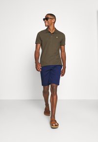 Mørk olivengrønn kortermet polo-skjorte med to-knappers placket, parret med marineblå shorts og brune sandaler. Tilbehøret inkluderer solbriller.