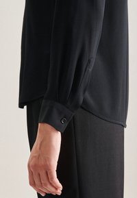 Maglietta nera a maniche lunghe con una texture liscia, caratterizzata da un polsino con bottoni e un orlo curvo, indossata sopra pantaloni neri.