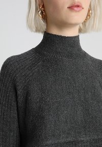 Ciemnoszary, prążkowany golf z wysokim kołnierzem i teksturowaną tkaniną. Zbliżenie pokazuje dodatek w postaci dużych kolczyków kołowych.