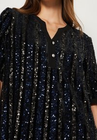 Svart paljettklänning med vertikala ränder i blått och silver, med knäppning fram och en V-ringning, korta ärmar och en texturerad yta.