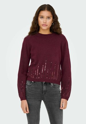 Bordeaux gebreide trui met paillettendetails aan de onderkant, ronde halslijn en lange mouwen, gecombineerd met donkergrijze jeans.