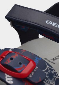 Κοντινό πλάνο παιδικού σανδαλιού σε σκούρο μπλε με κόκκινη αγκράφα, σχέδιο ιστού αράχνης και κείμενο, καθώς και ρυθμιζόμενο ιμάντα βέλκρο.