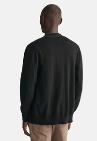 Schwarzer Pullover mit langen Ärmeln und gerippten Bündchen. Verfügt über einen runden Halsausschnitt und eine glatte Textur, von hinten gegen einen weißen Hintergrund betrachtet.