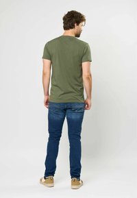 Alyvuogių žalia medvilnės džemperis, trumpomis rankovėmis, prigludęs dizainas, kartu su mėlynomis džinsinėmis kelnėmis ir smėlio spalvos sportbačiais neutraliame fone.