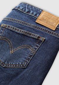 Jeans in denim blu scuro con una tasca posteriore caratterizzata da un motivo di cucitura distintivo e una toppa in pelle marrone chiaro con il marchio.