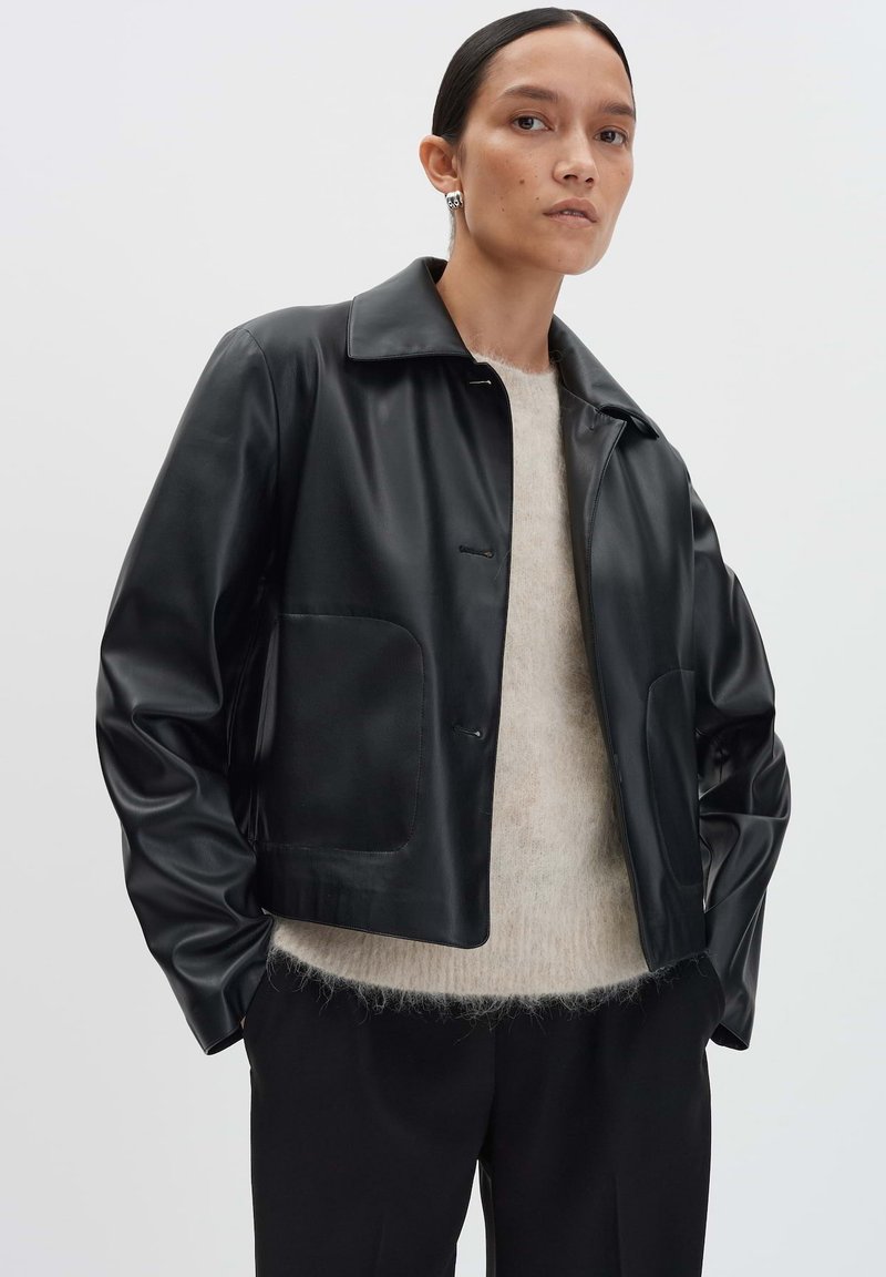 Giacca di pelle nera con collo, tasche frontali e una texture liscia, indossata sopra un maglione leggero e peloso e pantaloni neri.