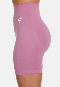 Zbliżenie na osobę w wysokich, bezszwowych szortach rowerowych w kolorze lawendowym z białym logo na lewym biodrze, na jednolitym tle.