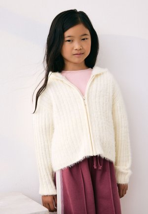 Maglione bianco a costine con zip e cappuccio, caratterizzato da una texture morbida. Indossato sopra un top rosa e pantaloni della tuta color bordeaux con coulisse.
