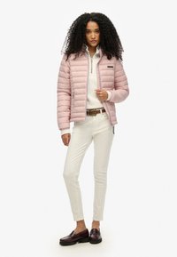 Jasnoróżowa kurtka puchowa z zamkiem z przodu, prążkowanymi mankietami i naszywką z logo. Nosi się ją na kremowym swetrze, w połączeniu z białymi spodniami i ciemnymi mokasynami.