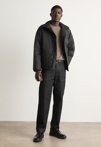 Giubbotto nero imbottito con cappuccio, maglione beige, pantaloni in denim scuro e scarpe nere. Il giubbotto ha polsini elastici e chiusura con zip.