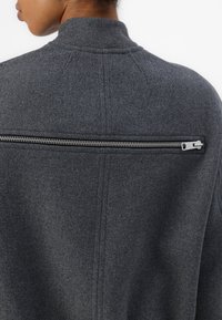 Grau gestrickte Jacke mit hohem Kragen und einer horizontalen Reißverschlusstasche über dem oberen Rücken, mit sauberen Nähten und einer strukturierten Oberfläche.