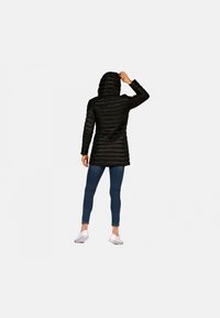 Veste noire matelassée avec une capuche, présentant un design en quilté horizontal et un dos légèrement plus long. Portée avec un jean bleu et des chaussures blanches.