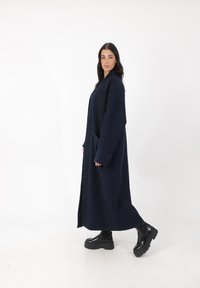 Abrigo largo azul marino hecho de tela gruesa con un corte relajado, que presenta mangas anchas y dos bolsillos prominentes. Combinado con botas negras de plataforma.