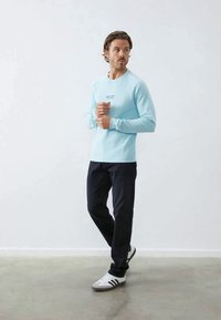 Svetlo modra majica z dolgimi rokavi z okroglim izrezom in majhnim logotipom, kombinirana s temnimi hlačami ter belimi športnimi čevlji s črnimi črtami.