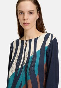 Långärmad topp i marinblått, krämfärgat och teal med djärva randmönster och nitar längs bröstet, med rund hals och nyckelhålDetalj.