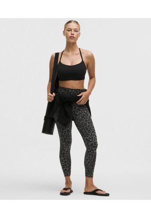 Frau steht und trägt einen schwarzen Sport-BH, graue Leggings mit Leopardenmuster, Flip-Flops, trägt eine Yogamatte in einer Tasche und hat eine schwarze Jacke um die Taille gebunden.