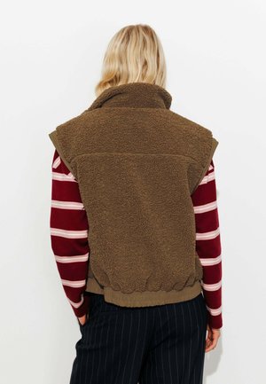 Veste sans manches en polaire marron avec col montant. Ourlet côtelé et détails de couleur contrastante au niveau du col. Portée par-dessus une chemise rayée.