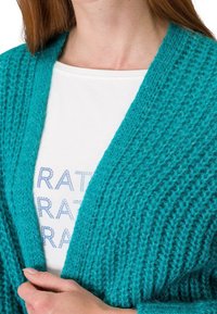 Türkiser Strickcardigan mit strukturiertem Muster, offener Vorderseite und geripptem Stoff. Über einem weißen Hemd mit bedrucktem Text getragen.