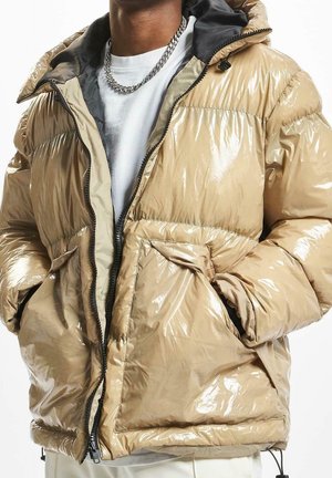 Person trägt beige glänzende Steppjacke mit Kapuze, weißes Hemd, silberne Kettenhalskette, Hände in den Jackentaschen, vor weißem Hintergrund.