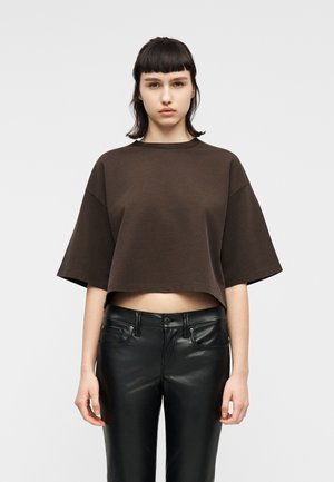 Mladá osoba s krátkými tmavými vlasy, která má na sobě volné hnědé krátké tričko a černé kožené kalhoty, stojící před jednoduchým světlým pozadím.