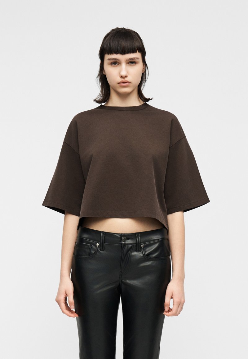 Νεαρό άτομο με κοντά σκούρα μαλλιά, που φοράει φαρδιά καφέ κοντή μπλούζα και μαύρο δερμάτινο παντελόνι, που στέκεται μπροστά σε ένα απλό φωτεινό φόντο.