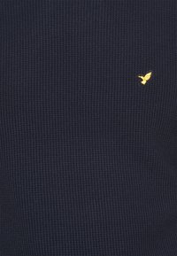 Pulover din tricot cu model texturat, de culoare bleumarin. Include un mic logo de pasăre aurie pe piept, evidențiind un design simplu și modern.