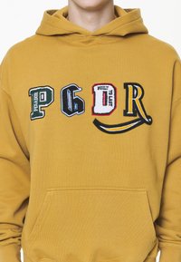 Felpa gialla con un grande tascone frontale, caratterizzata da lettere e dettagli ricamati multicolore che riportano la scritta "PEGADOR BUILT TO LAST."
